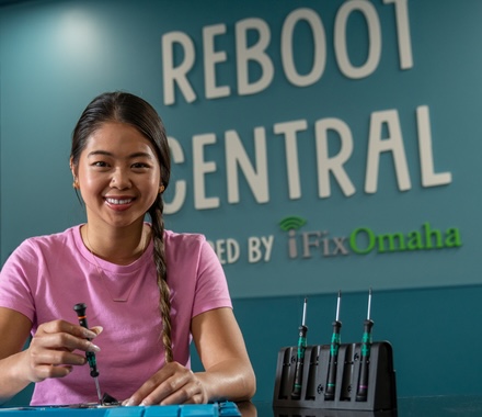 A female student with long black hair wears a pink shirt while repairing a smartphone with a screwdriver at the Digital Express "Reboot Central" desk.
