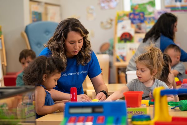 A woman early childhood education professional works assists two young girls sitting at a table engaged in a group classroom project in a childcare setting.